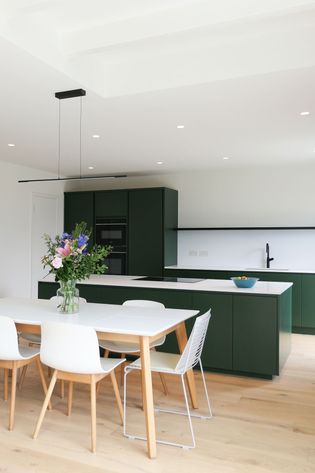 Modern kitchen with green cabinets, white countertops, and light wood flooring. A dining table with flowers adds a fresh, elegant touch.