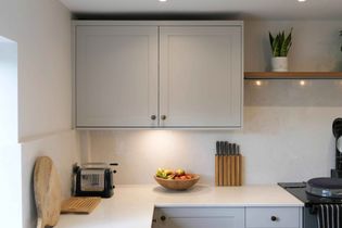 Modern kitchen with white cabinets, a bowl of fruit, knife block, and toaster on the counter. A small plant sits on a shelf, creating a clean, cozy feel.