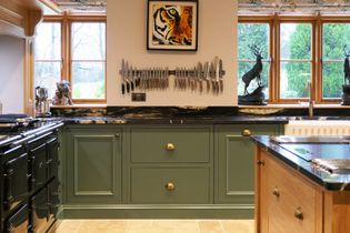 Stylish kitchen with green cabinets, black countertop, and neatly arranged knives on a wall. Warm lighting, window view, and a decorative deer statue.