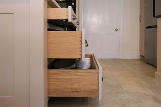 Open wooden kitchen drawers showing neatly stacked baking pans and dishes. The scene conveys a tidy, organized kitchen with light-colored tiles and cabinetry.