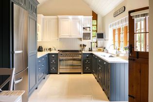 Shaker kitchen with blue lower cabinets, white upper cabinets, and a stainless steel fridge. Sunlight filters through large windows, creating a cozy atmosphere.
