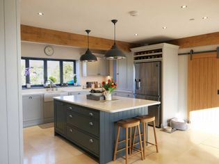 Shaker kitchen with a central island, wooden stools, pendant lights, grey cabinets, and a stainless steel fridge. Bright, inviting atmosphere.
