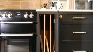 A shaker kitchen with a black stove featuring silver knobs. To the right, dark cabinets with gold handles; a cabinet door open reveals vertical dividers. A quartz worktop above holds utensils, creating a sleek and organized atmosphere.