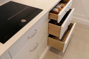 Shaker kitchen with three open white drawers containing utensils and white plates, next to a sleek black stovetop on a light countertop. Clean and organized.
