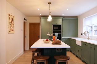 Shaker kitchen with light green cabinets and contrasting dark green central island. A loaf of bread and fruit bowl sit on the island. Warm lighting and wooden accents throughout.