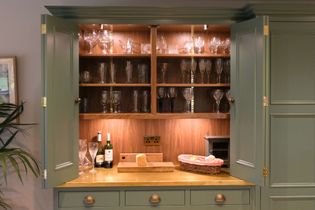 Open green cabinet with glassware, wine bottles, and a cheese board on a wooden counter. Warm lighting creates a cozy, inviting atmosphere.