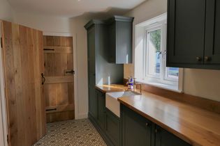 A utility with dark cabinets, wooden countertops, and a farmhouse sink. Sunlight streams through a window, illuminating the patterned floor tiles.