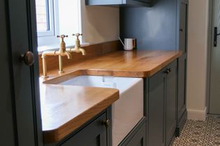 A shaker utility with dark cabinets, wooden countertops, and brass taps. A white farmhouse sink and patterned tile floor add a chic touch.