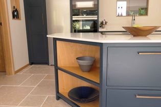 Shaker kitchen with a blue island featuring a white countertop and wooden shelves. Decor includes ceramic bowls, a fruit bowl, and visible appliances.