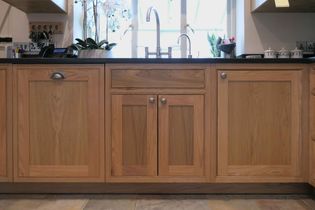 Wooden kitchen cabinets with silver handles below a black countertop and silver sink, set against a sunlit window. Clean and organized.