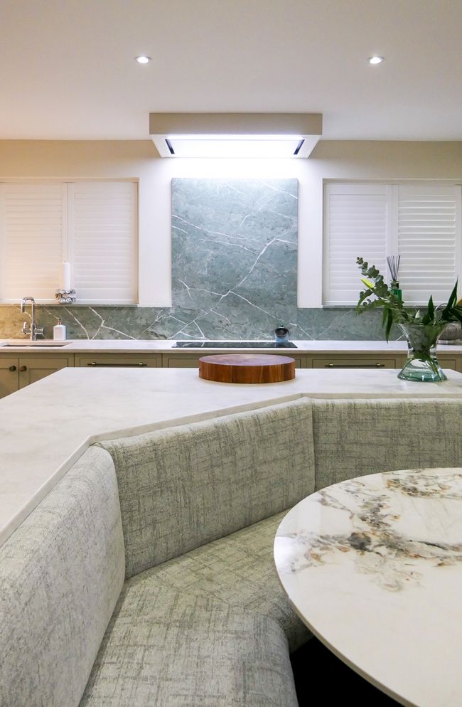 A Shaker kitchen featuring a light-coloured countertop, a round table, and cosy bench seating area with a grey fabric cushion.