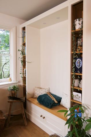 Shaker style window nook with a wooden bench, cushions, and a throw. Shelves with plants, books, and decor with natural light streaming in.