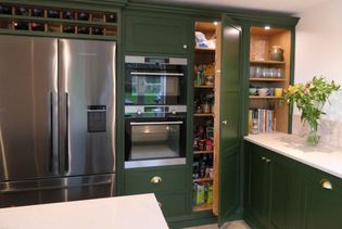 Shaker kitchen with green cabinets, stainless steel fridge, built-in oven, open pantry with food items, and a countertop with flowers and books.