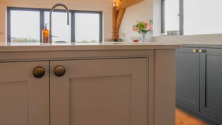 Shaker kitchen with a white island, brass fixtures, and dark cabinets. A vase of pink flowers and fruit bowl sit by a window, creating a cozy feel.