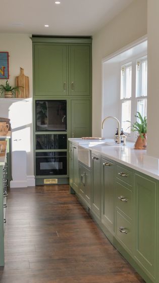 Shaker cabinetry run with two ovens at the end, an open front farmhouse sink is in the middle of the cabinetry with a green plant on the window sill.