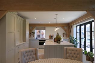 Shaker kitchen with cream cabinetry, white island, and black-framed windows. It features pendant lights, plants, and a comforting dining area with upholstered chairs.