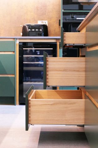 A modern kitchen with green cabinetry, featuring open wooden drawers, and a black refrigerator in the background.