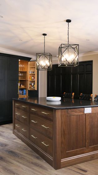 Shaker kitchen with dark wooden island, black countertop, and brass handles. Two geometric pendant lights hang above. Open pantry shelves reveal snacks.