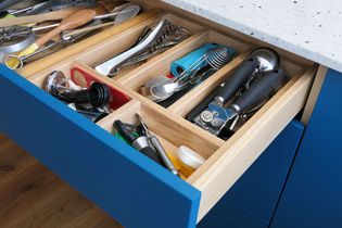 Open kitchen drawer with neatly organized utensils. Wooden dividers separate items like can openers, ice cream scoops, and other gadgets. Blue exterior and light countertop.