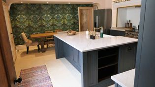 Spacious kitchen with island, dark cabinets, and ornate green wallpaper. Cozy dining area with a wooden table and cushioned chairs in the background.