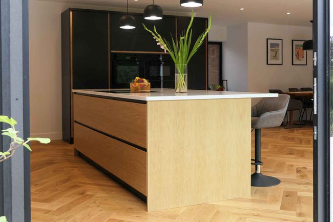 Modern kitchen with a wooden island countertop, pendant lights, flower vase, and bar stools on herringbone wood flooring.