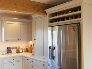 Shaker kitchen with light grey cabinets and a stainless steel refrigerator. Wooden beam above adds warmth. Wine bottles are stored above the fridge.