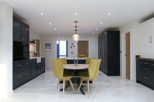 Shaker kitchen with a round dining table and 4 mustard-yellow chairs on light tile flooring. Dark grey cabinetry contrasts with white panels. Overhead lighting adds warmth.