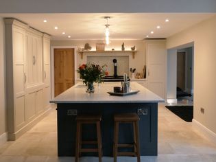 Shaker kitchen with a large island, two wooden stools, overhead pendant lighting, and cream cabinets. A vase of flowers adds warmth and charm.