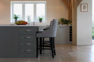 Shaker kitchen with a dark grey island, high-back grey bar stools, and a large window. Decor includes fruit, potted plants, and elegant wooden beams.The perimeter cabinetry is a light grey colour.