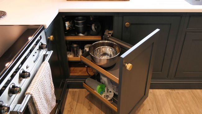A shaker kitchen features dark cabinets with brass knobs. A pull-out corner cabinet reveals mixing bowls, appliances, and utensils. The stove has a towel draped over the handle.
