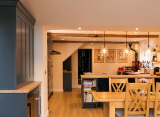 Warmly lit open-plan kitchen and dining area with exposed beams, pendant lights, dark cabinetry, and a wooden dining table.
