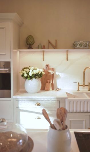 A Shaker kitchen with white cabinetry, a bouquet of white flowers in a round vase, wooden cutting boards, and a gold faucet above a sink.