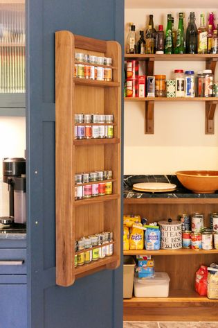 A wooden spice rack mounted on a Shaker door, filled with various spice jars, next to a kitchen area with shelves displaying pantry items.
