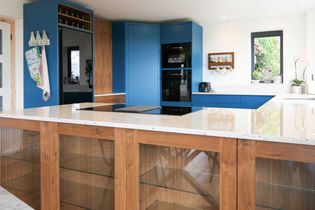 Modern kitchen with blue cabinets, wooden accents, and a white countertop. Features a glass cabinet, wall oven, and garden view window.