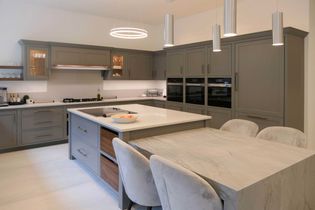 Shaker kitchen with grey cabinets, a large marble island with a bowl of fruit, sleek pendant lights, and built-in double ovens. Bright, clean ambiance.