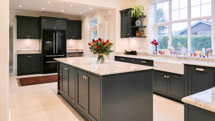 Bright large Shaker kitchen with dark cabinetry and white worktops, the central island has a vase of flowers in the middle, a farmhouse sink below the window with various items on the windows sill and open shelving.