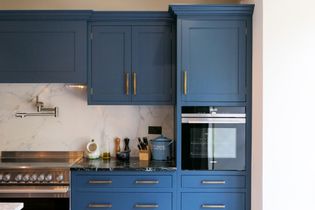 A Shaker kitchen featuring blue cabinets, a stainless steel oven, a black countertop, and marble backsplash, with various kitchen utensils on display.