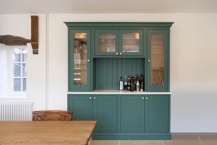 A green dresser with glass doors, displaying bottles and glasses, is set against a white wall. A wooden table and chair are partially visible. Cozy and tidy ambiance.