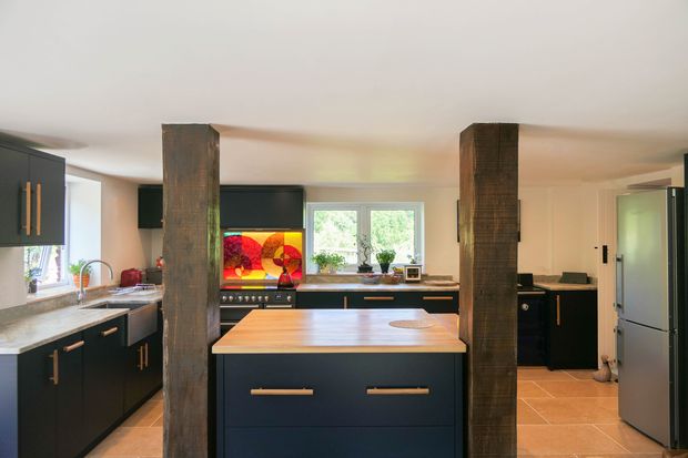 Modern dark kitchen with a central island, two rustic wooden posts, colourful abstract artwork, and windows overlooking greenery.