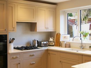 Shaker kitchen with natural cabinets, light countertops, and a stainless steel gas stove. Large window with potted plants overlooks a backyard.