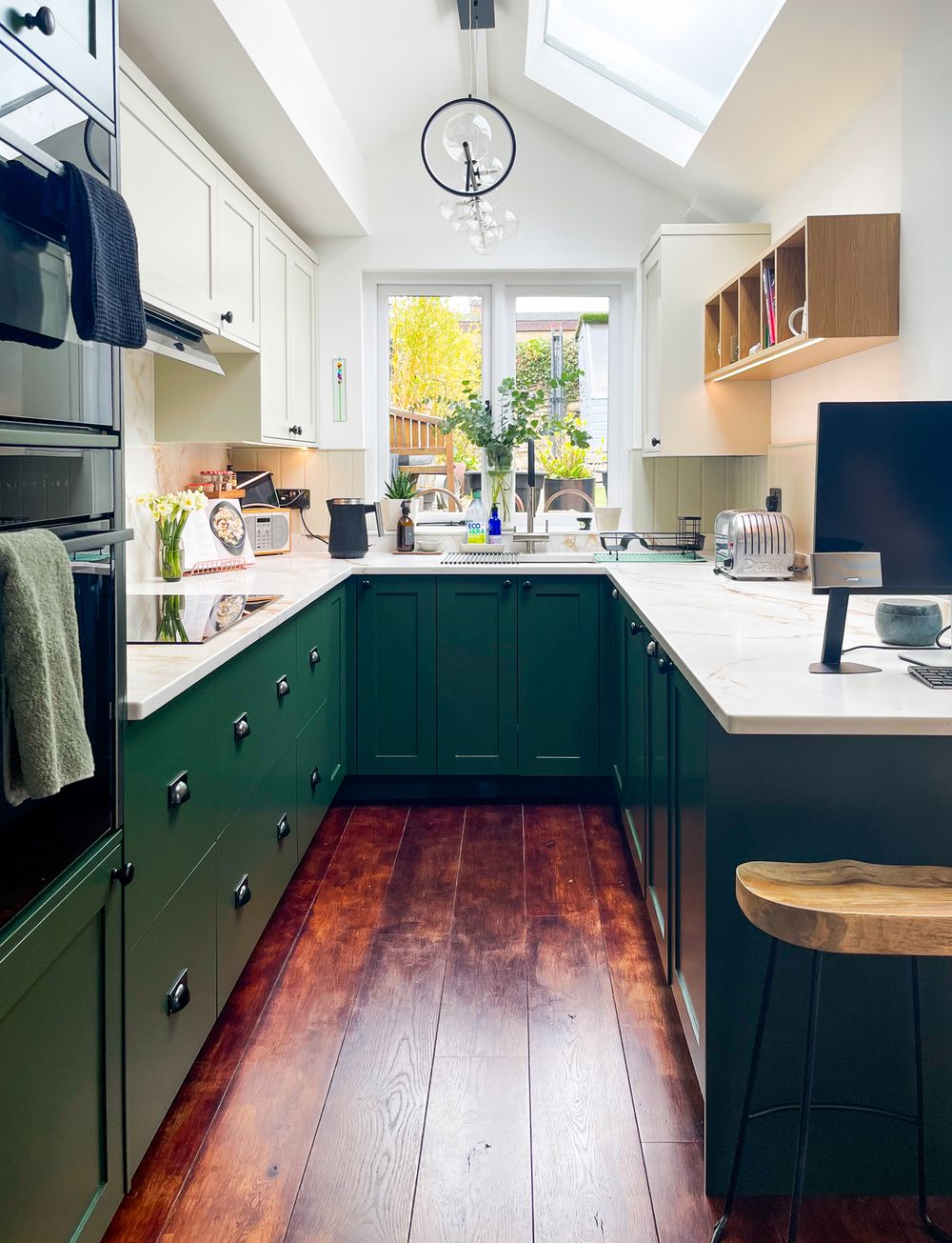 Bright u-shape Shaker kitchen with dark green cabinets, white countertops, skylights, and a garden visible through the central window.