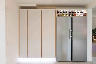 Minimalist kitchen with sleek white cabinetry and a stainless steel double-door fridge. A lit shelf above displays assorted bottles, creating a modern ambiance.