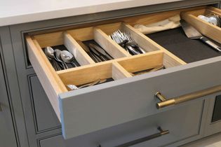 Open kitchen drawer with organized wooden cutlery tray. Spoons, forks, and knives neatly arranged. Gray drawer with brass handles, conveying tidiness.