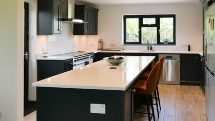 Shaker kitchen with dark cabinets, a large white island, orange bar stools, stainless-steel appliances, and a window overlooking greenery.