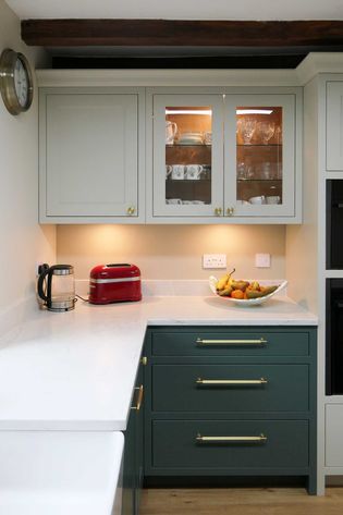 Shaker style kitchen with neutral and green cabinets. A countertop holds a red toaster, kettle, and fruit bowl. Warm lighting inside and underneath the cabinets. 