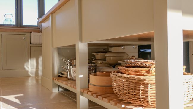 A Shaker kitchen with a wooden island, featuring shelves underneath that hold various pots, pans, and a wicker basket.