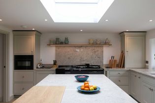 Bright kitchen with soft grey cabinets, a large marble island, and a skylight. Fresh fruit and bowls on the counter add a vibrant touch. Cozy and inviting.