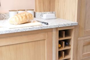 A kitchen counter with wooden oak cabinetry, featuring a loaf of bread in plastic wrap on a cutting board, a knife, a butter dish, and wine storage.