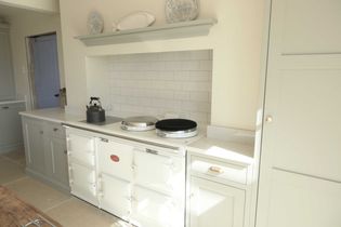 A bright kitchen corner features a AGA white stove with a black kettle, set against a subway tile backsplash and soft cream cabinets. Plates adorn a shelf above.