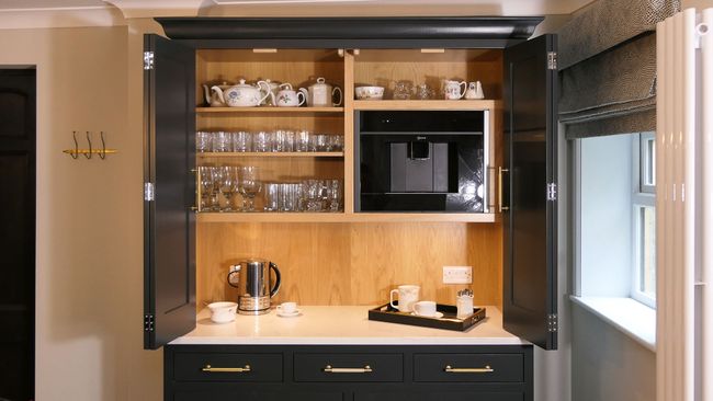 Open kitchen cabinet with dark doors reveals neatly arranged shelves of teapots, glasses, and a built-in coffee maker; countertop holds a kettle and tray.